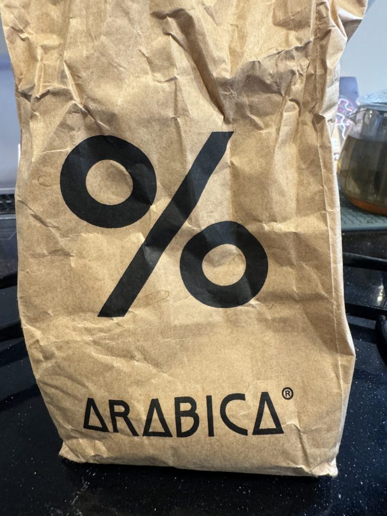 A brown paper bag with the "% Arabica" logo printed on it, sitting on a black surface.